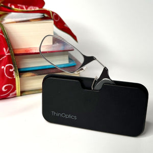 A pair of ThinOptics Readers popping out of a Universal Pod Case presented next to a pile of books.